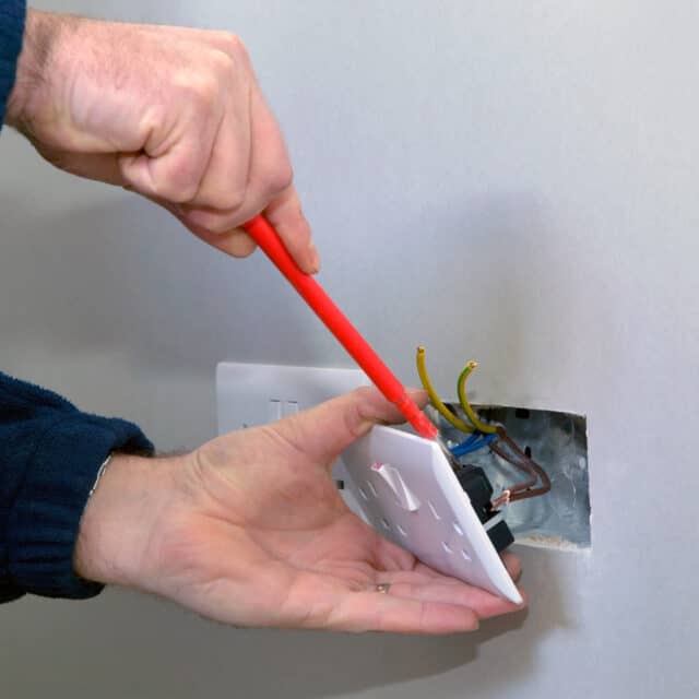 Person using a red screwdriver to install or repair a wall electrical outlet, with exposed wires visible inside the wall cutout.
