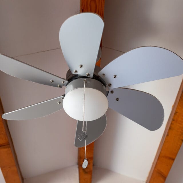 Ceiling fan with five alternating gray and white blades, a central light fixture, and pull chains, mounted on a ceiling with exposed wooden beams.
