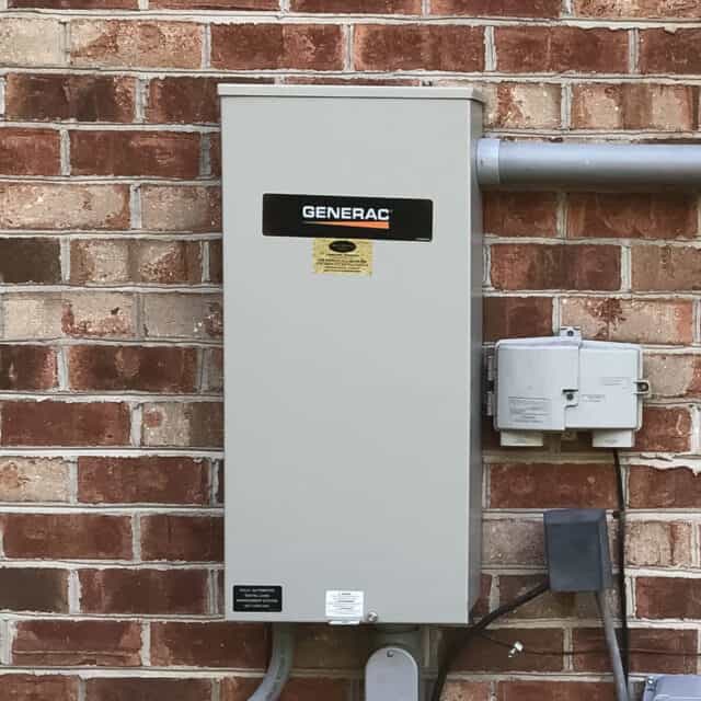 Outdoor Generac electrical panel mounted on a brick wall, with conduits and nearby utility boxes.