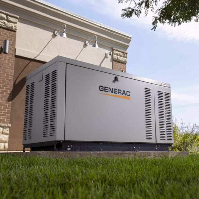 Large Generac generator on a concrete pad beside a building, surrounded by grass, with trees partially visible above.
