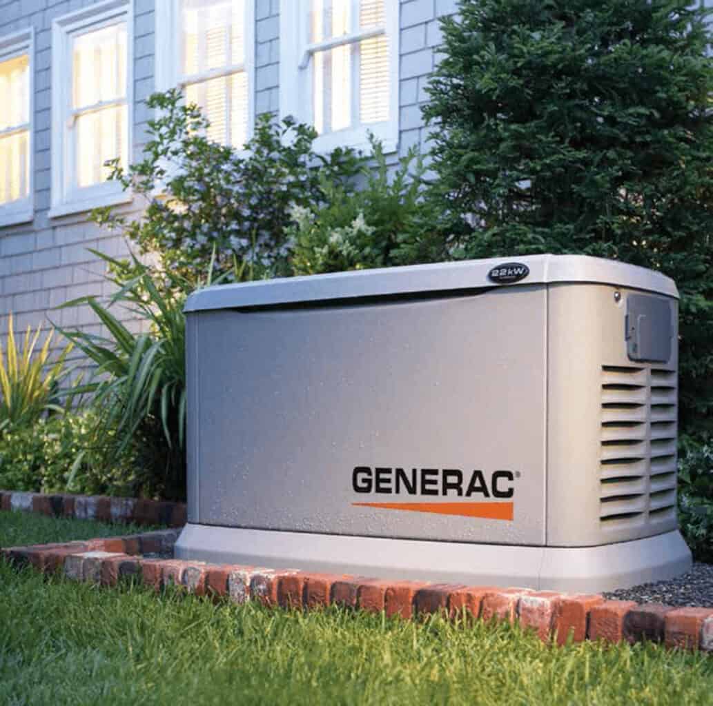 A Generac generator installed outside a house on a brick platform, surrounded by grass and small shrubs.