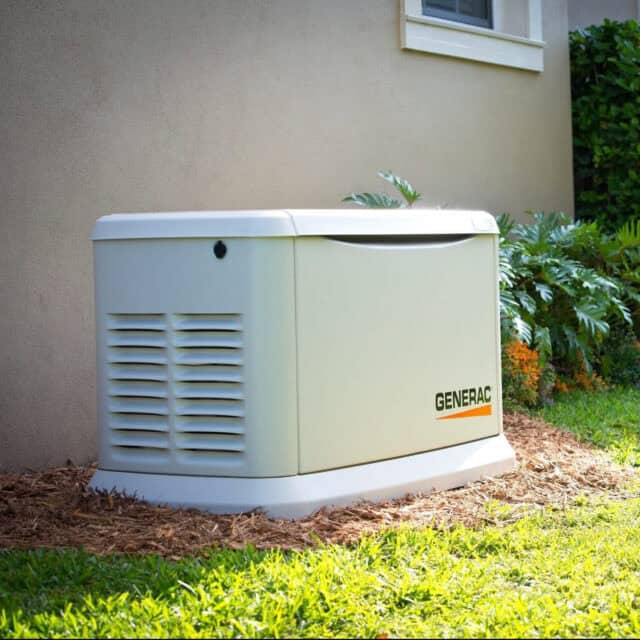 A Generac standby generator is installed next to a house, surrounded by mulch, with green grass and plants nearby.